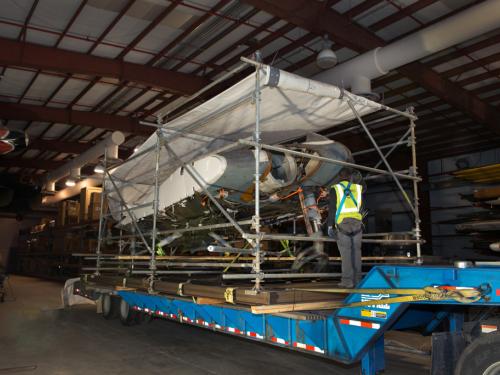 A gray and white metal aircraft, attached to a steel support structure, sits on the long and large bed of a transportation truck. A white tarp protects the top of the aircraft.