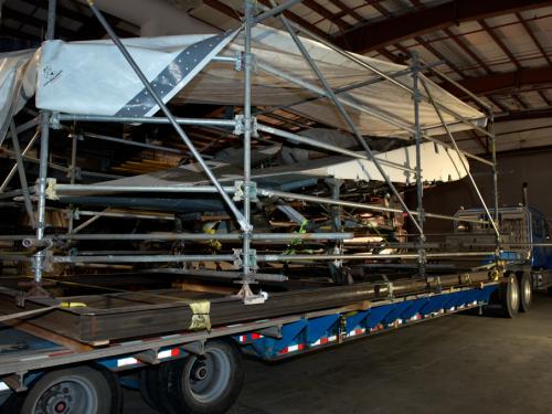 Diagonal back view of the Horton Ho 229 V3, a gray and white metal aircraft, loaded onto a truck for transport. The top of the aircraft is covered by a large white tarp for protection,