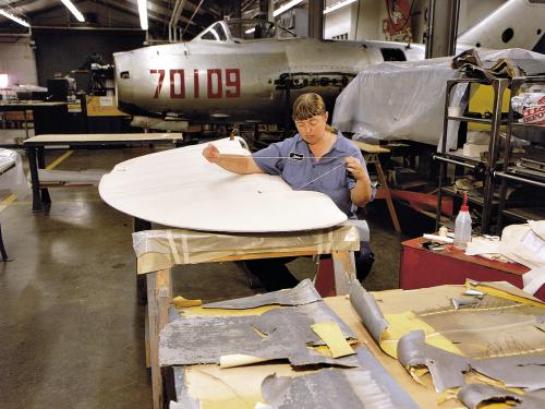 Restoration staff member stitching a control surface is seated holding a line of thread over a beige fabric-covered piece of an aircraft.