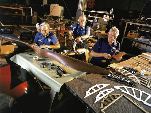 Three restoration volunteers are seated and smiling while working on aircraft parts. A volunteer in the foreground holds sandpaper over the center of a large wood propellor. 