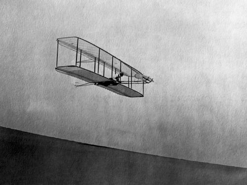 A biplane gliding in the air down a hill. A person lays on top of the bottom wing, perpendicular to it, as they pilot the craft.