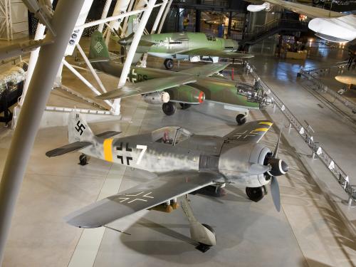 Three German, military aircraft from World War II are displayed next to each other at the Udvar-Hazy Center. The closest aircraft is a silver-colored aircraft and the two aircraft behind it are olive drab green.