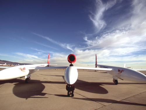 Front view of wide, low winged white aircraft sitting on tarmac with blue sky in the background.