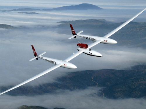 View from above of wide white, low-winged aircraft in flight over cloud-topped mountains.