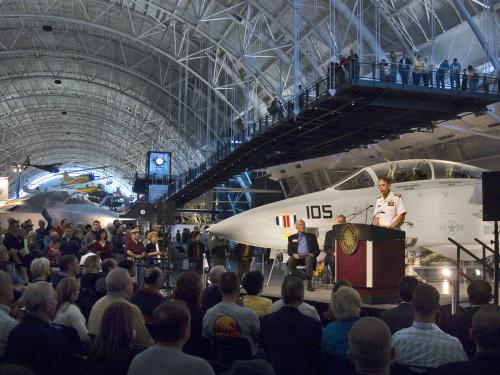 A former director of the National Air and Space Museum speaks in front of a crowd as he welcomes a white fighter jet located behind him at the Udvar-Hazy Center.