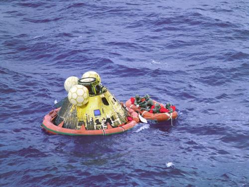 An asymetrical, yellow-colored cylindrical prism sits in a float next to its crew in an orange raft prior to being recovered following the Apollo 11 mission.