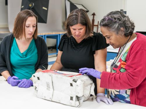 Three female Museum staff members, one of whom is a conservator and the other two are curators, look at a white and red oxygen purge system cover used on the Moon that was recently unsealed.