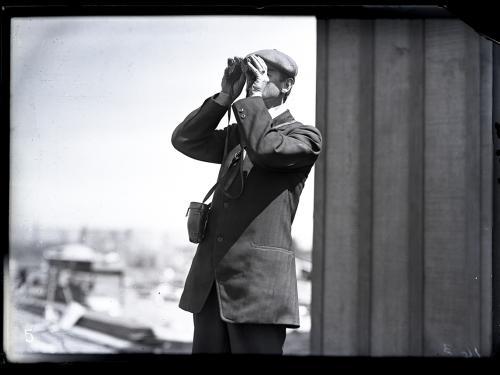 A man looks through binoculars with a column behind him. 