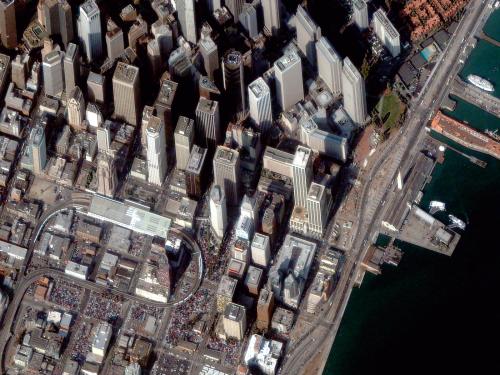 Close-up, satellite view of San Francisco, with high rise buildings to the north and smaller buildings and the San Francisco-Oakland Bay Bridge to the south.