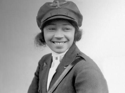 Bessie Coleman, a woman who appears to be in her late 20s, smiles at something to the right of the camera. She is wearing a military-like uniform, with an emblem that reads "B.C." on her cap.