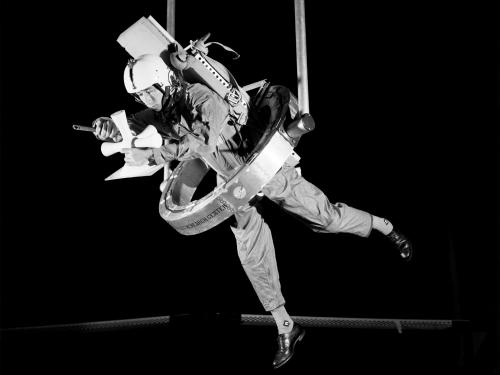 In this black and white image, a man is suspended from an apparatus while using his hands to handle a device. 