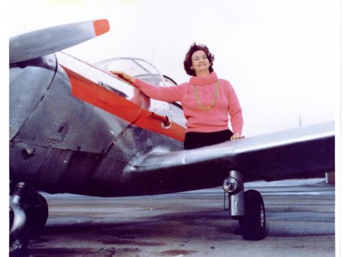 A partial view of the left side of an airplane with a white then red stripe under the cockpit. A woman in a pink mock turtleneck sweater with a gold chain necklace sits on the wing of the airplane. She has lost both legs above the knee.