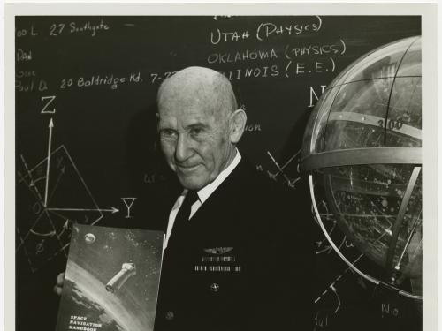 Bald man in Navy uniform stands in front of a blackboard holding a book. Blackboard with chalk writing in the background, Celestial navigation globe partially visible at right.