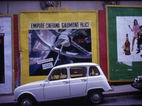 A white vehicle is parked in front of a movie poster for the film "2001: A Space Odyssey" plastered on multicolored wall. 