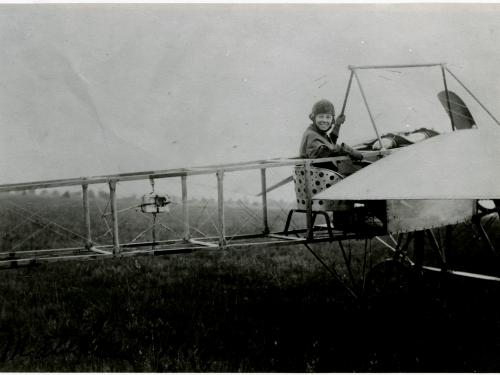 Rearview shot of an early airplane with Moisant in cockpit looking over her shoulder and smiling at the camera. 