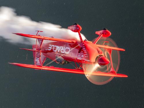 Red biplane flies inverted over dark water, smoke plume coming off the top