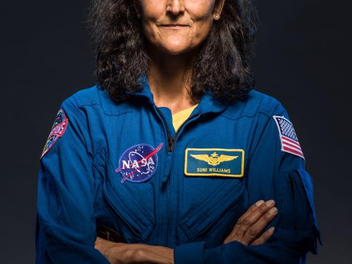 NASA Astronaut Sunita Williams looks at the camera, with a dark background, in a blue suit. 