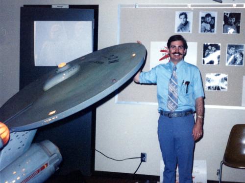 A white male poses next to a gray spaceship studio model from Star Trek.