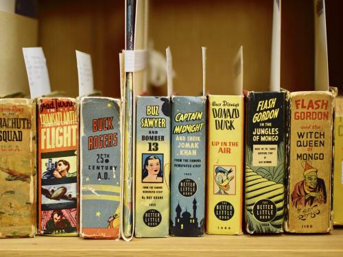 A row of short squat books with index cards peeking out. The books stand up with the spines facing out. Each spine is colorful and boasts a comic like "Flash Gordon," Buck Rodgers," or "Donald Duck Up in the Air."