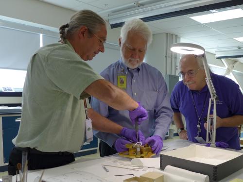 Three people stand around the film transport and work on it on a table. 