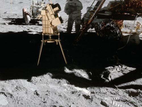 Image of camera on the Moon; Lunar Module, Astronaut, and American flag in background. 