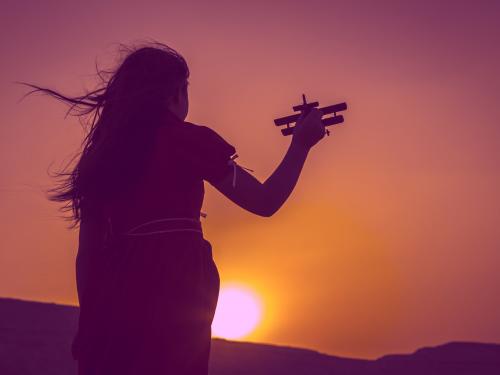 Image of young girl holding up an airplane against a backdrop of the sunset. 