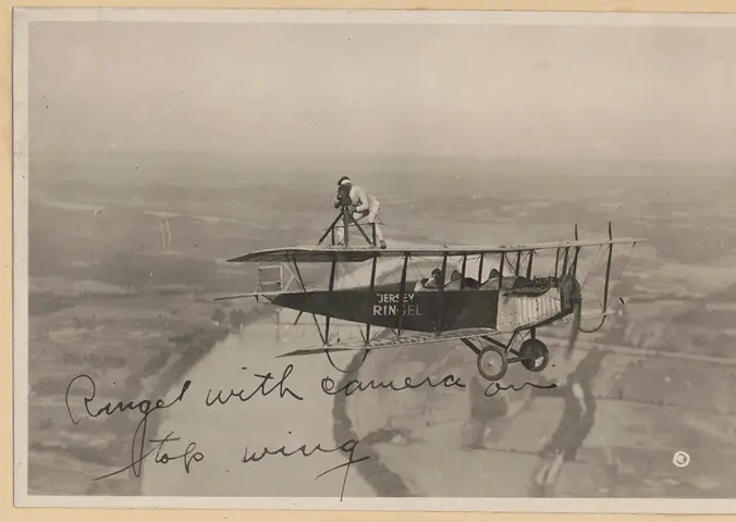 A man performing a barnstorming act standing on the wing of an airplane holding a camera