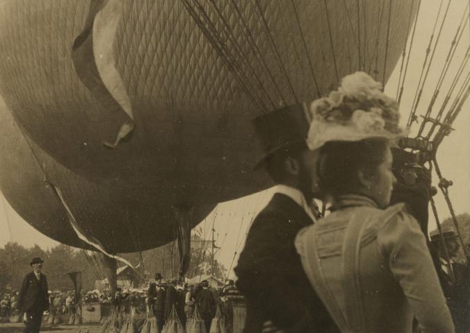 A sepia tone photograph, in the foreground are fashionably dressed people from the early 1900s, in the background are hot air balloons. 