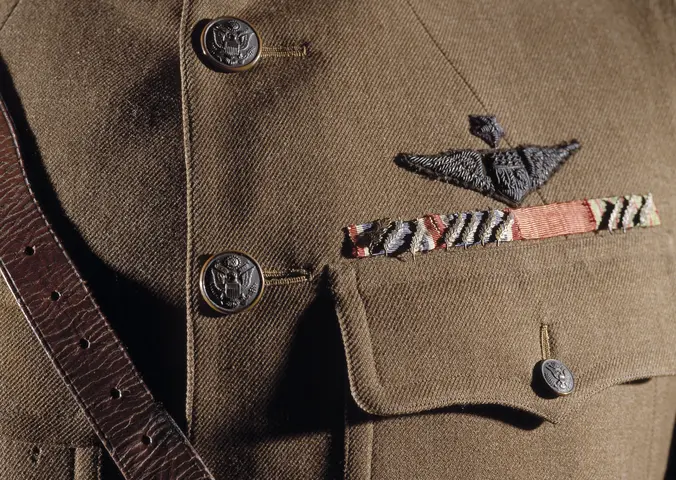A single breasted drab wool coat with five brass buttons down front; two upper patch pockets and two lower patch pockets with brass buttoned flap; a stiff or "choker" collar; a single gold officer's lace on each cuff; a brass "U.S." insignia on right collar; a Winged Propeller Air Service insignia on left collar; three overseas service chevrons on left cuff; a Distinguished Service Cross ribbon with 7 oak leaf clusters, a French Legion of Honor ribbon, and a French Croix de Guerre ribbon with 3 palm leaves.