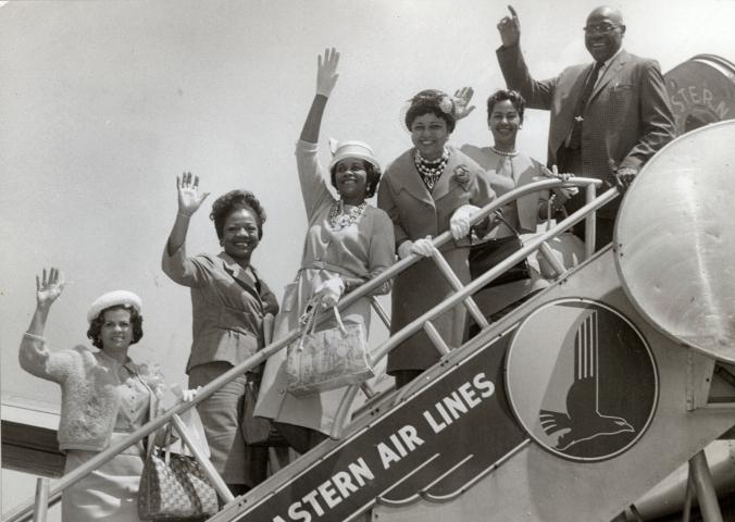 A group of African-American people pose and wave on the boarding steps of an aircraft.