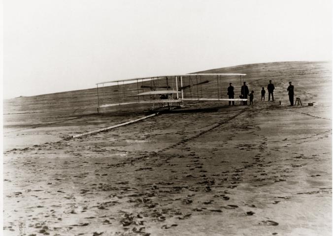 A biplane sits on a long wooden rail on the ground. A group of people stand behind it.