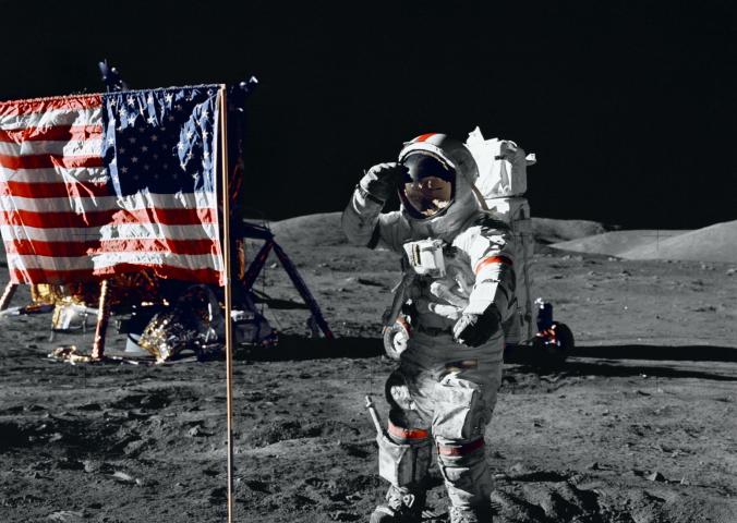 Eugene A. Cernan, a male astronaut wearing a full-body spacesuit, salutes a red, white, and blue U.S. Flag on the surface of the Moon.