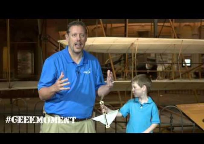Marty Kelsey stands in front of the Wright Flyer with a small child playing with a toy.