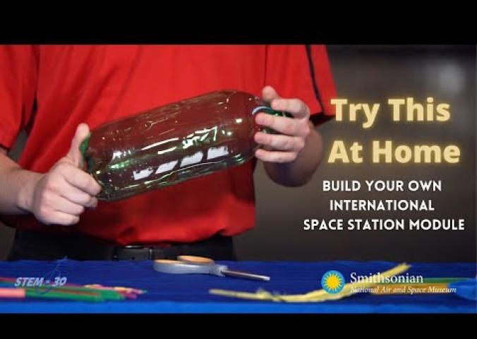 A video of an Explainer discussing how to build a space station with materials from home! The Explainer is in a red polo and stands behind a table with items on it.