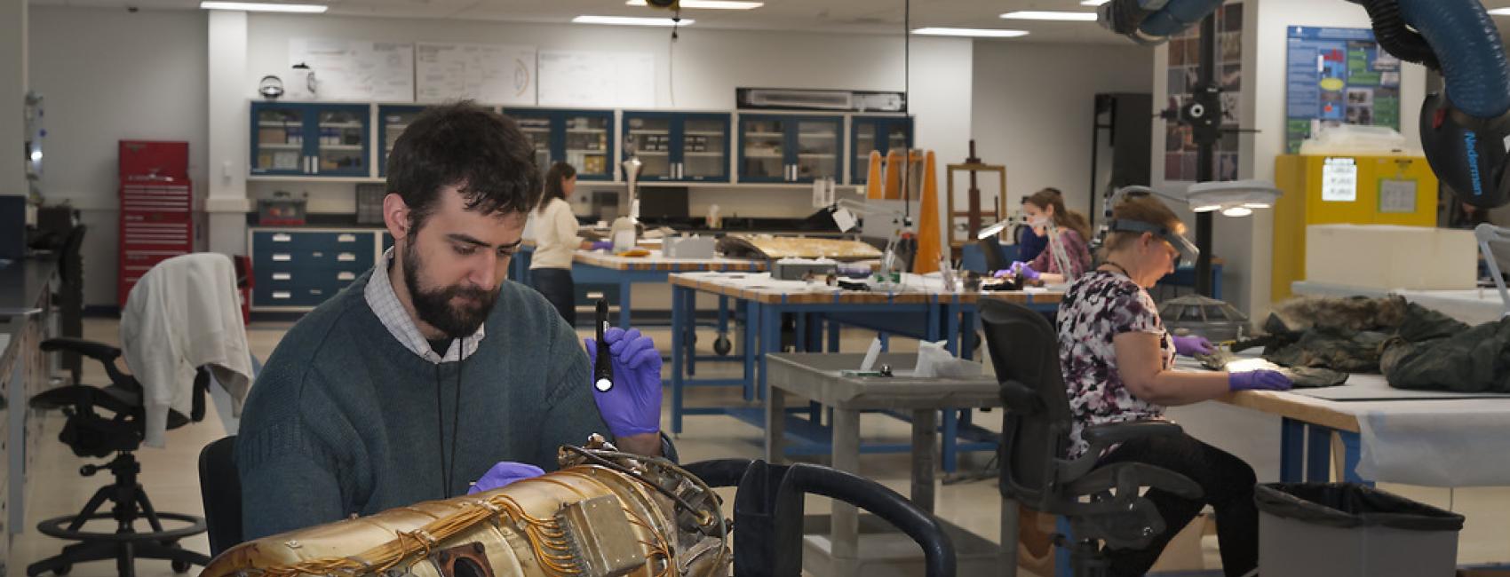 People at work in the conservation lab. In the foreground, a man holds a flashlight over a bronze cylinder with wires on it. In the background women sit at tables, the objects they're working on cannot be seen. 