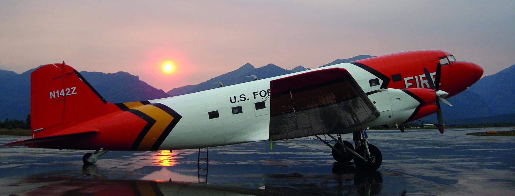 A DC-3 parked on the tarmac with a setting sun in the distance. The plane is red, orange, and white, and has the word fire across the front part of the plane.