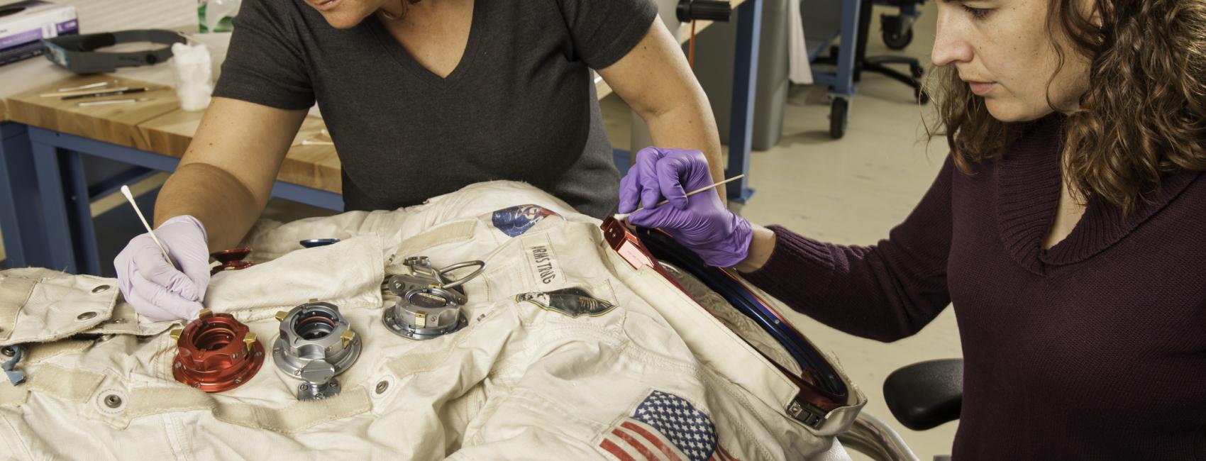 Two women next to a spacesuit working to conserve it with tools.