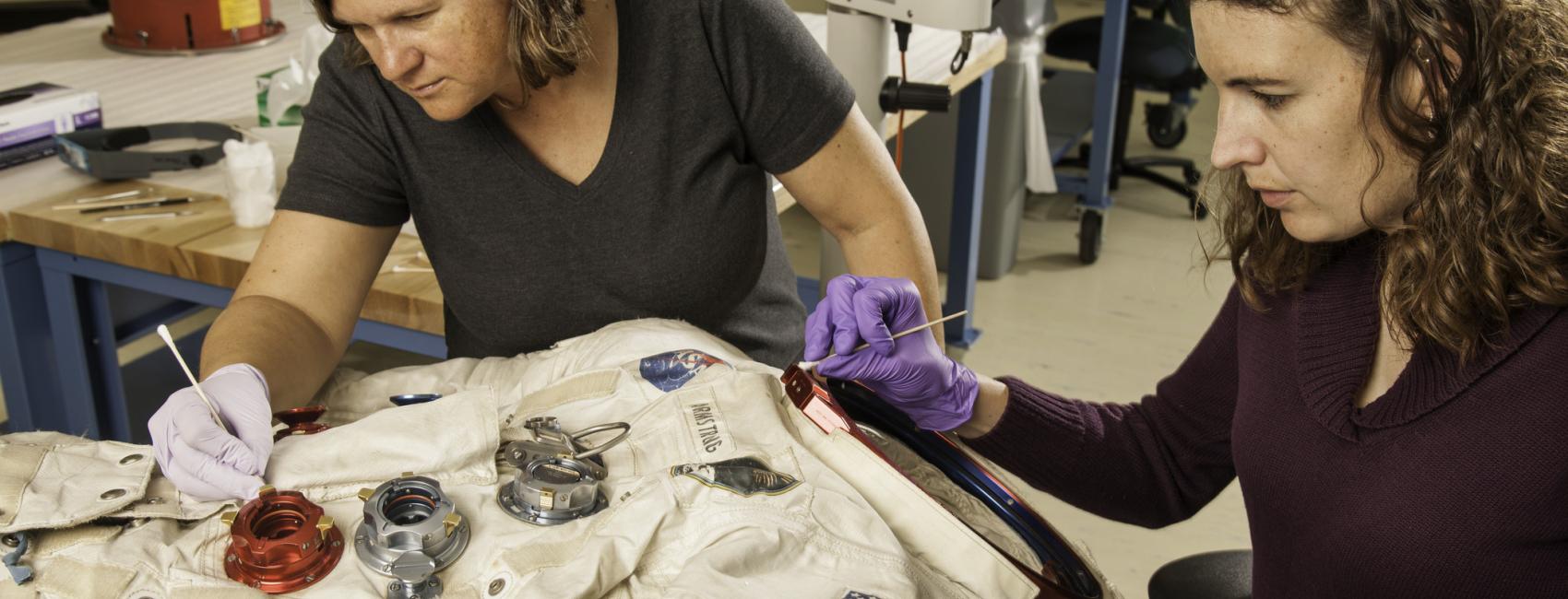 Two women next to a spacesuit working to conserve it with tools.