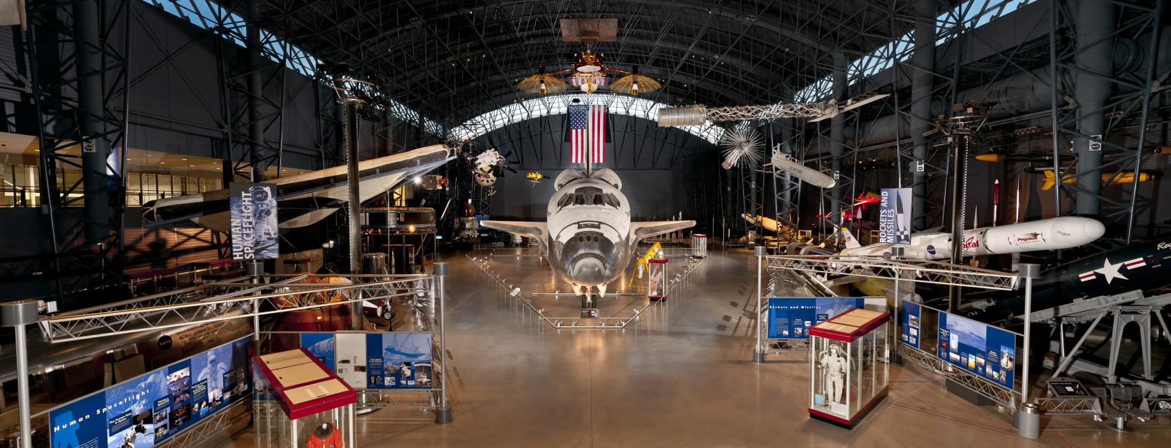 At the center of the photograph is the nose of the Space Shuttle, with the Space Shuttle expanding behind it. Hanging overhead is a satellite, a spacesuit, and a missile. Surrounding on the ground are cases with space stuff. 