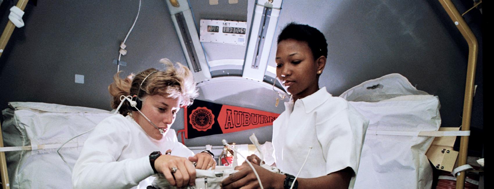 Two women floating in zero gravity work look at a scientific instrument. 