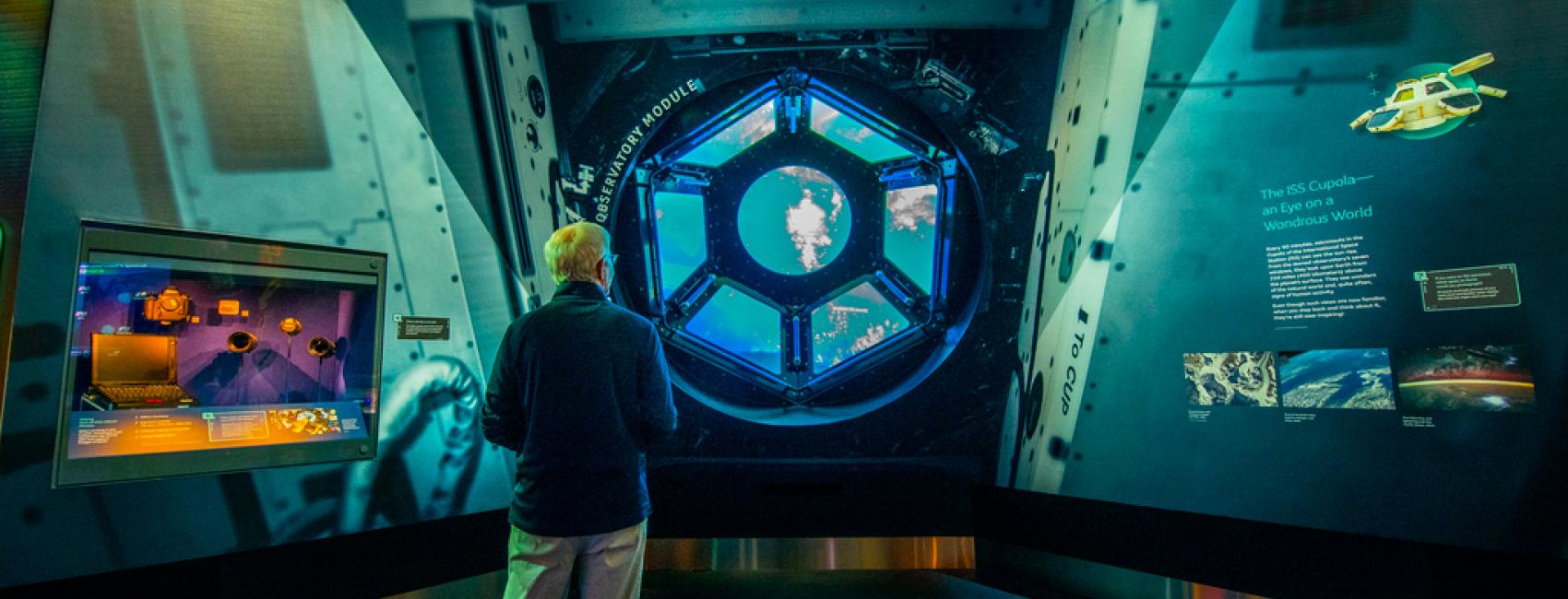 A person with white hair looks out a replica of the cupola window on the International Space Station. Through the window a video recording of Earth from space is visible. 