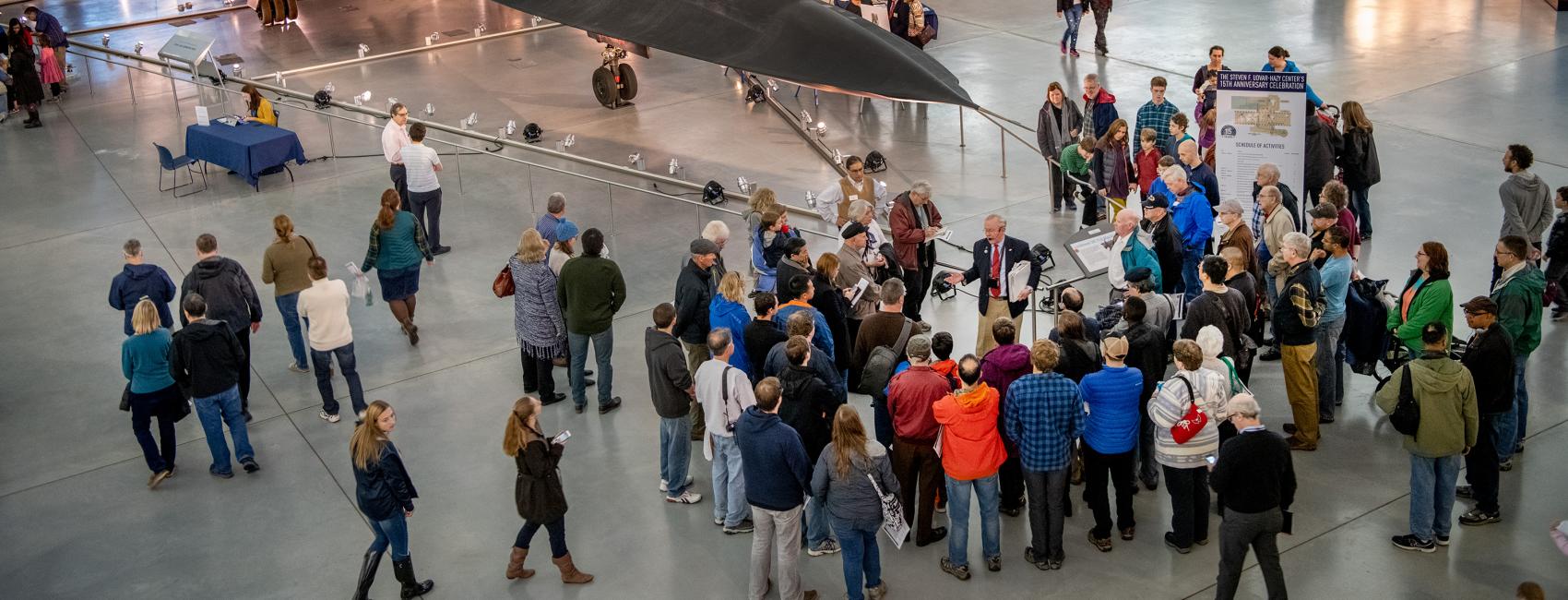 A group of people stand by the nose of the SR-71, a big black plane. One person is speaking to the group.