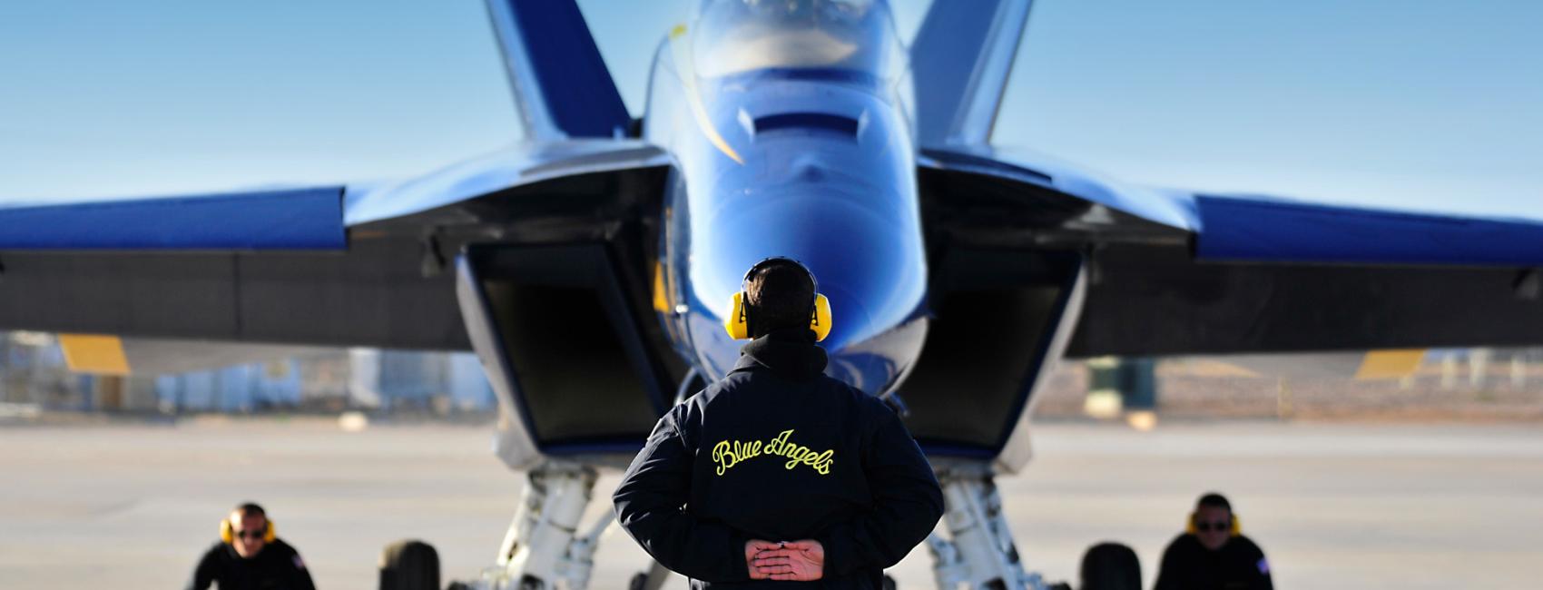 A person wearing a "Blue Angels" jacked and yellow ear protection stands up straight, with their hands locked behind their back and their back to the camera. In front of them, in the background of the photo is a Blue Angel plane and two people crouched down under it. You are led to believe the plane is about to take off. 