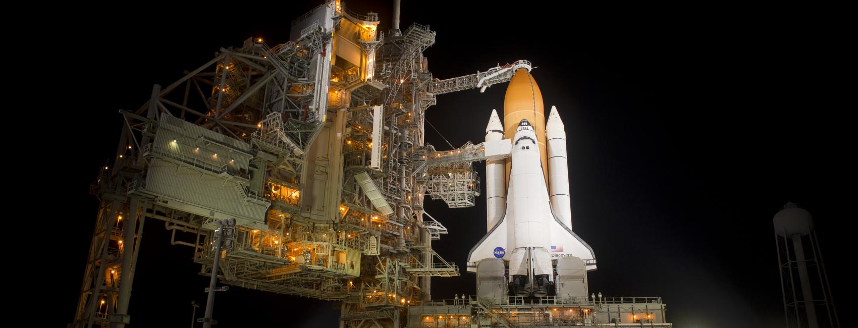 A space shuttle on the launchpad, with its nose pointed toward the sky. The night sky makes up the background.