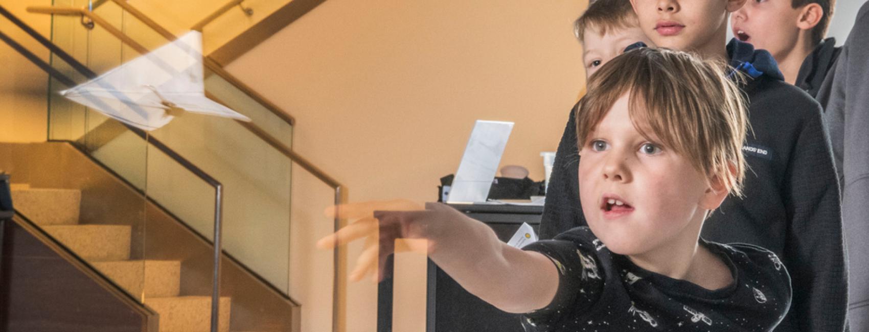 A young visitor is rapt with attention as they throw a paper airplane, with a similar line of young visitors behind them. 