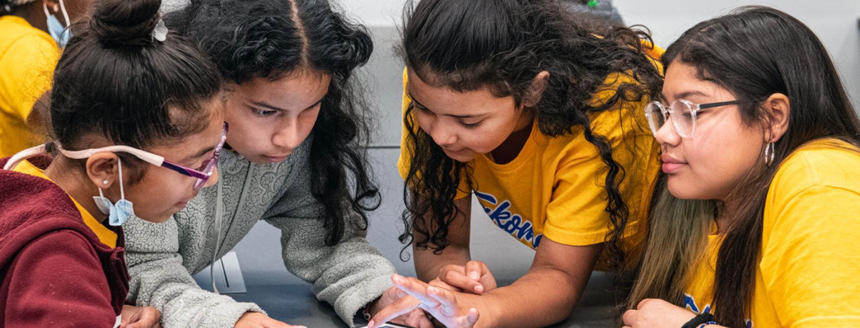 Four young girls hunch over a tablet, working on a problem. 