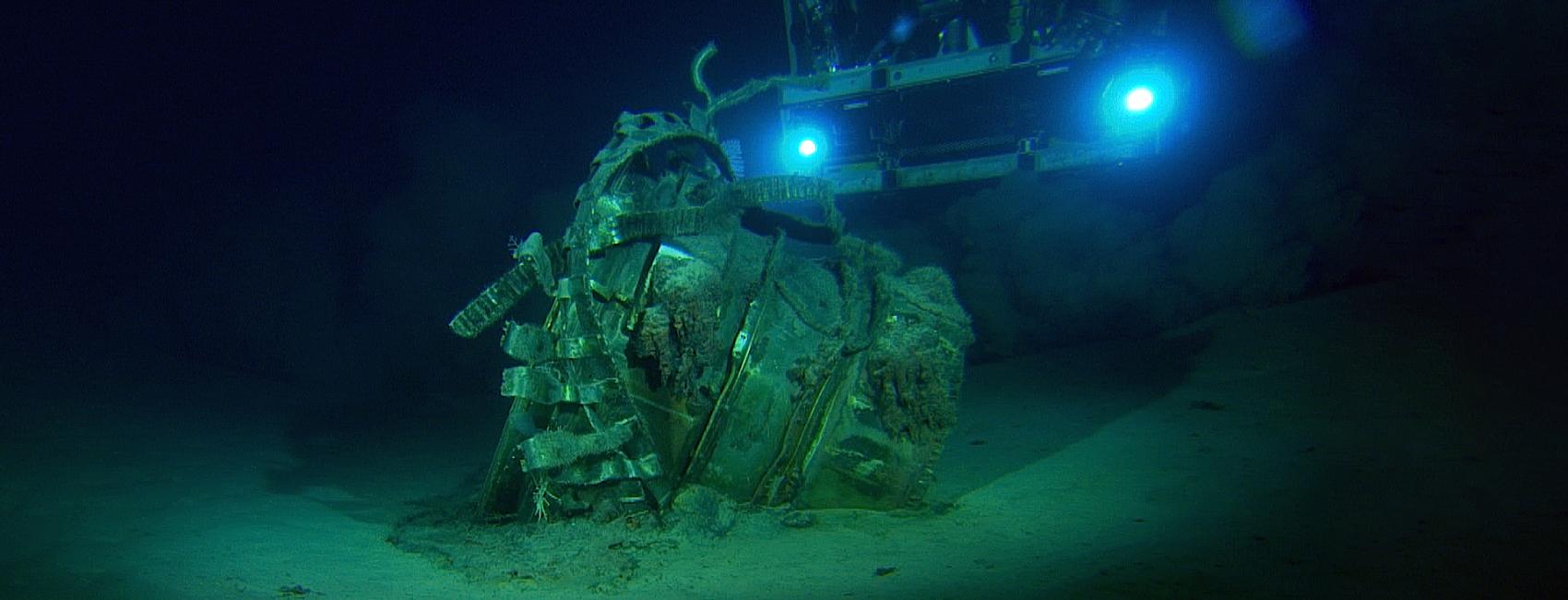 Underwater image of a large metal object, resting on the ocean floor. The object is partially covered with marine growth, indicating it has been submerged for some time. In the background, an underwater vehicle equipped with lights illuminates the scene.