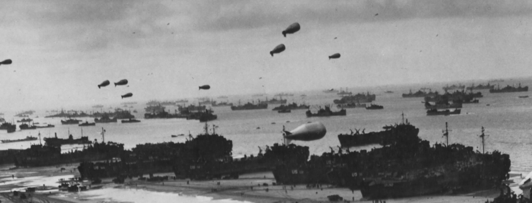 A horizontal black and white photo of balloons floating above a beach covered in military gear. 