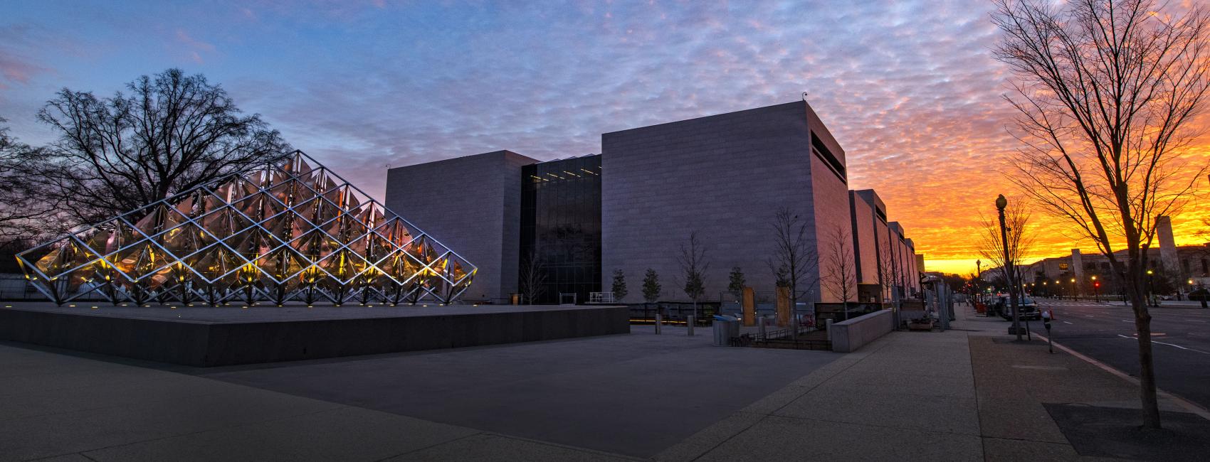 The sunset paints the sky a rainbow ombre outside of the National Air and Space Museum. A triangular metal sculpture stands silhouetted against the multicolor sky. 