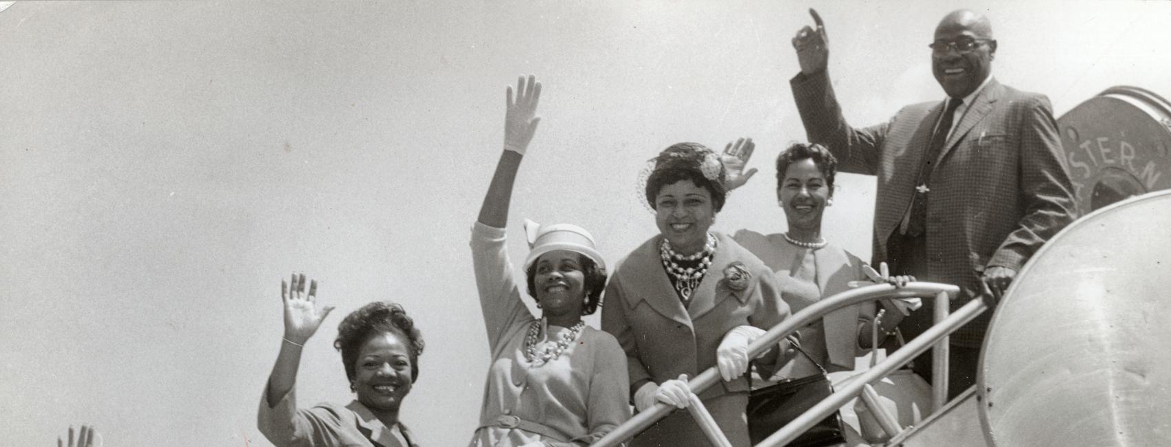 A group of African-American people pose and wave on the boarding steps of an aircraft.